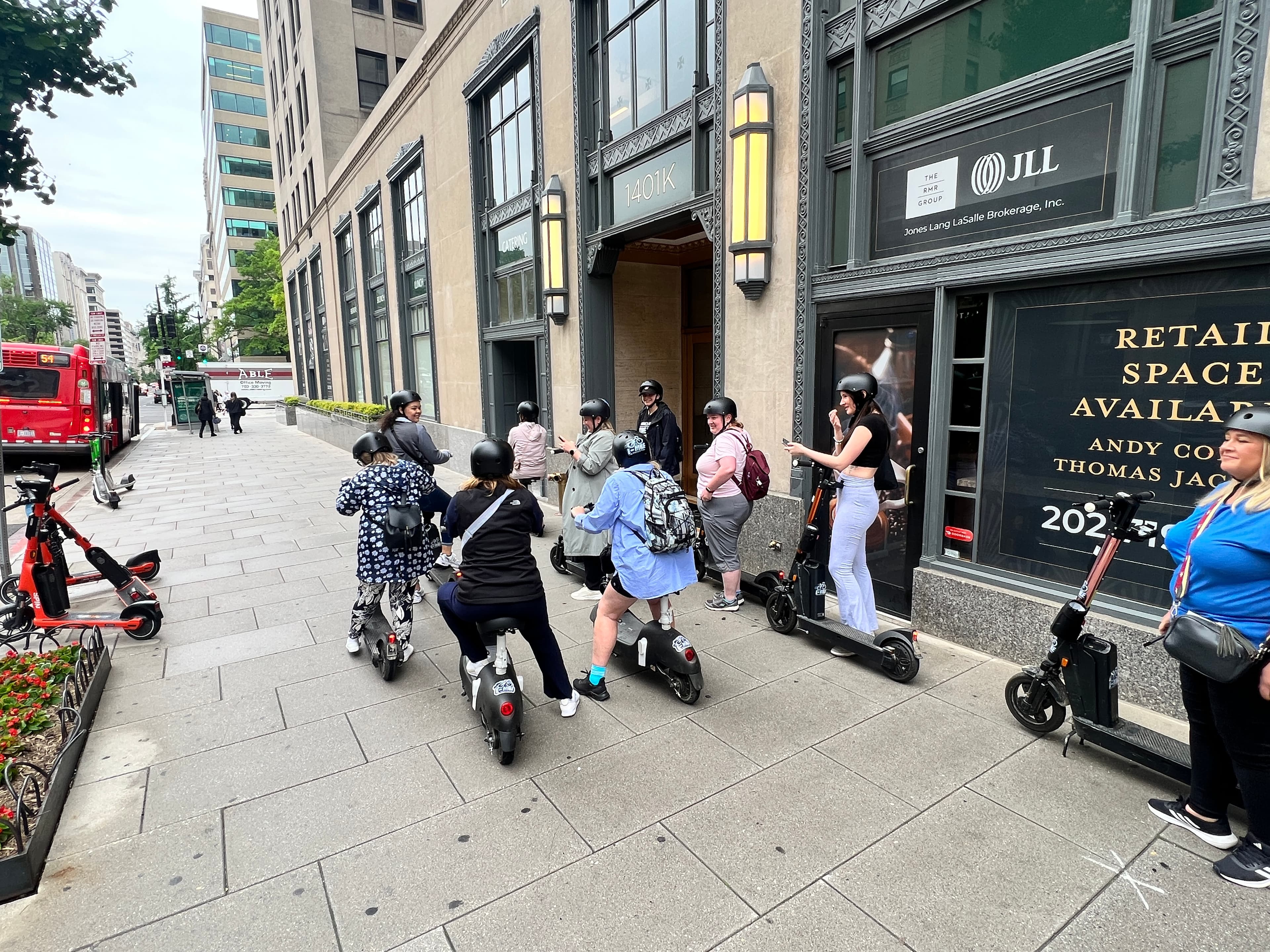 Group of people wearing helmets on electric and seated scooters on a city sidewalk.