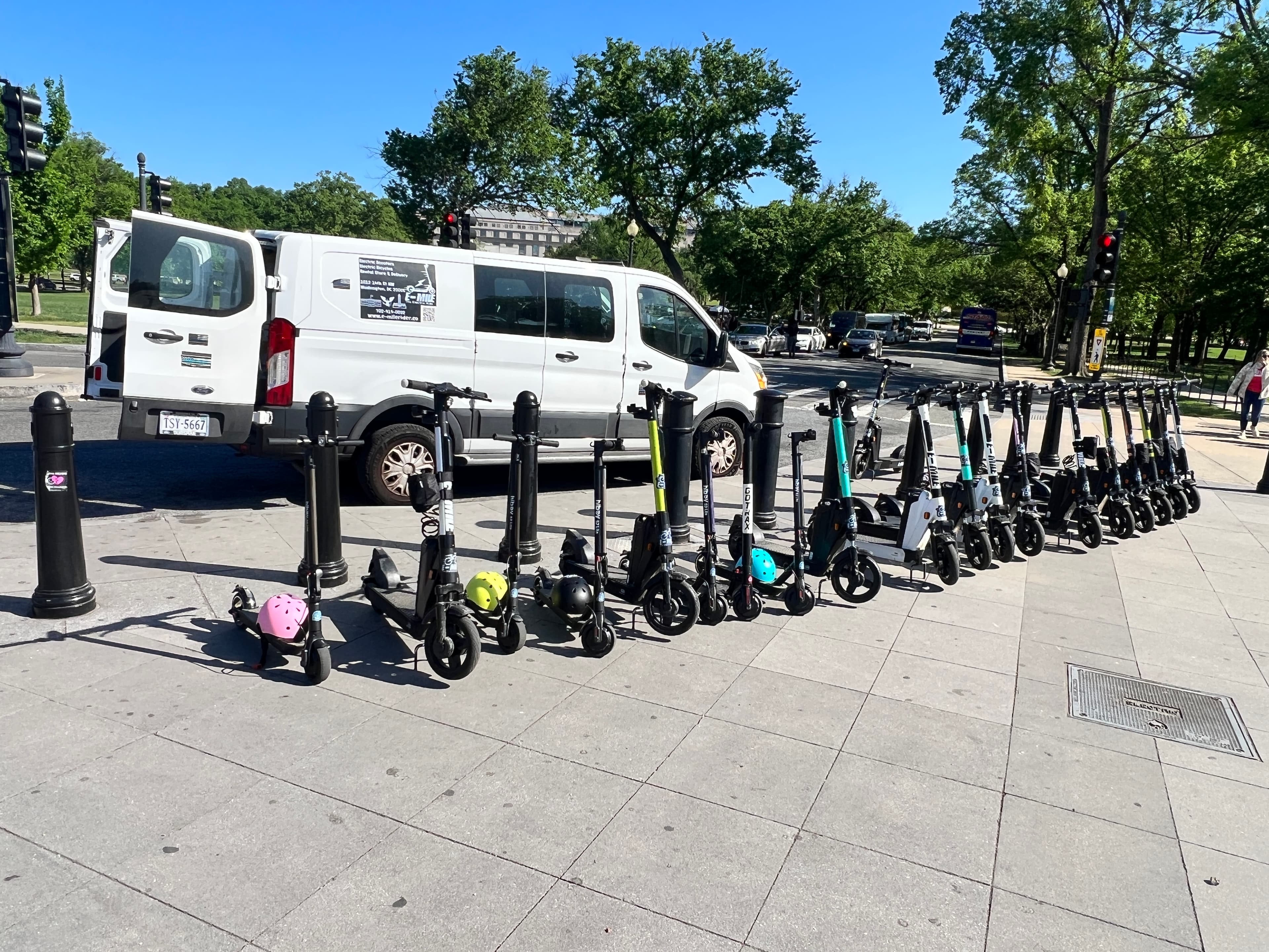 Electric scooters with colorful helmets parked on a city sidewalk next to a white van.