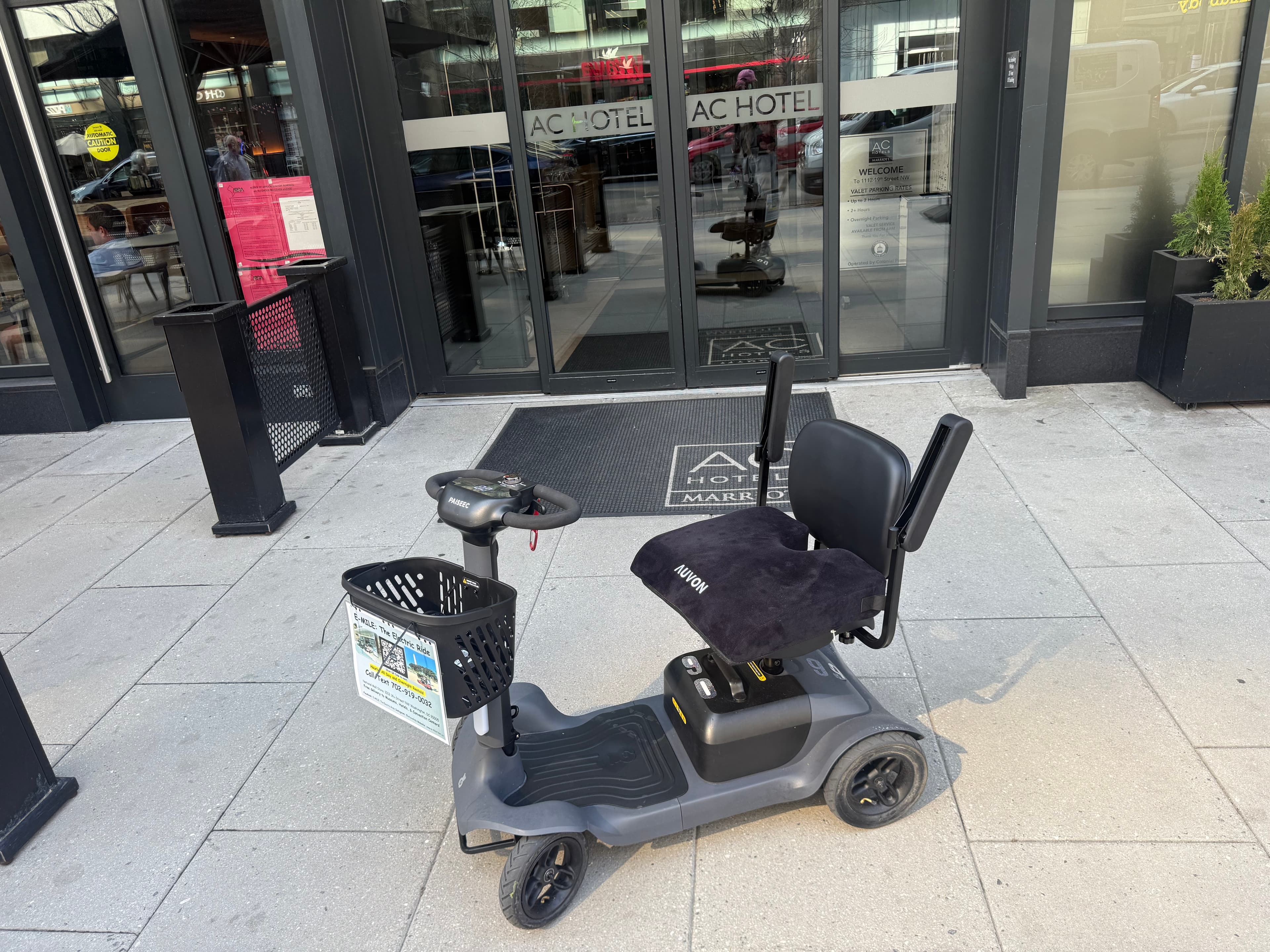 Grey mobility scooter with a black seat cushion parked outside an AC Hotel entrance.