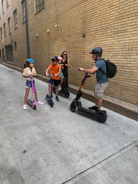 Adults and children wearing helmets stand on electric scooters in a narrow brick alleyway.