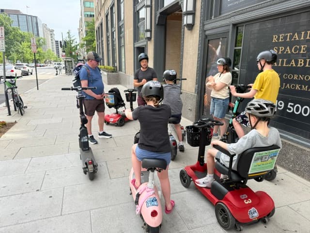 A group on electric scooters and mobility devices listens to a guide on a city sidewalk.