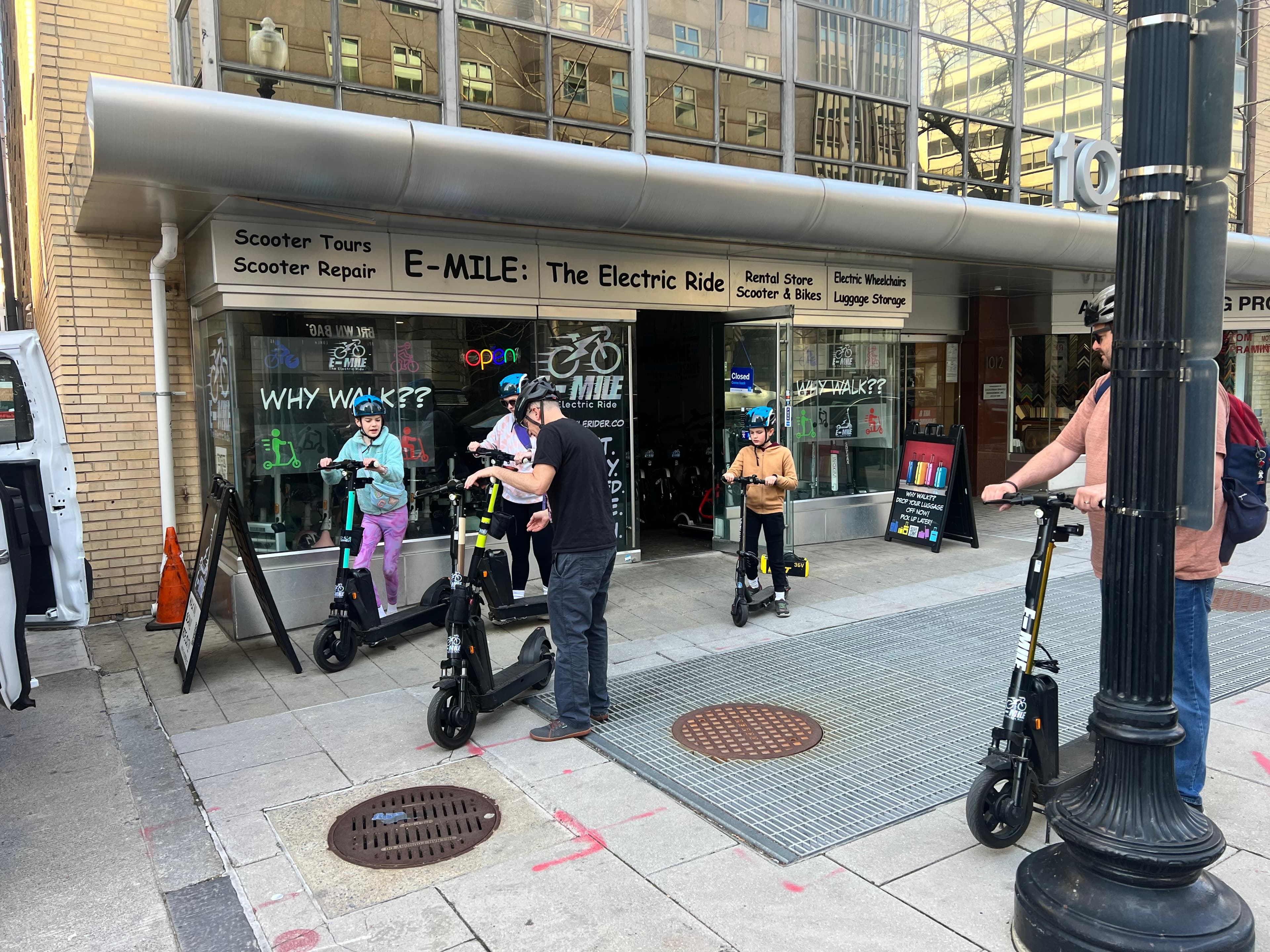 People prepare to ride electric scooters outside an E-MILE rental shop on a city sidewalk.