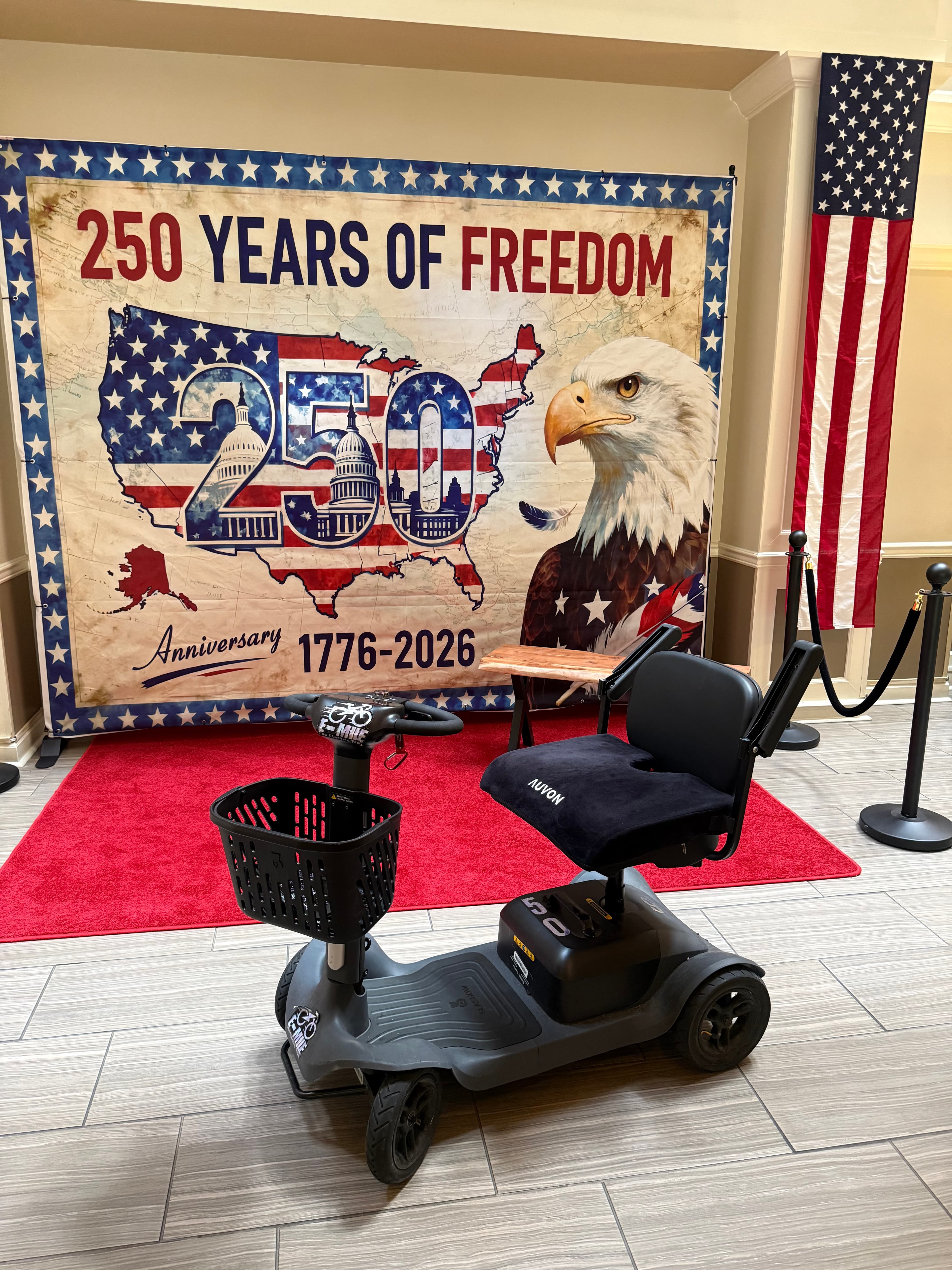 Black mobility scooter on a red carpet in front of a patriotic 250th anniversary banner.