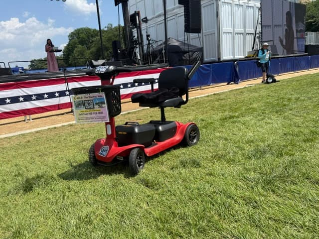 Red mobility scooter with a basket parked on grass in front of an outdoor stage.