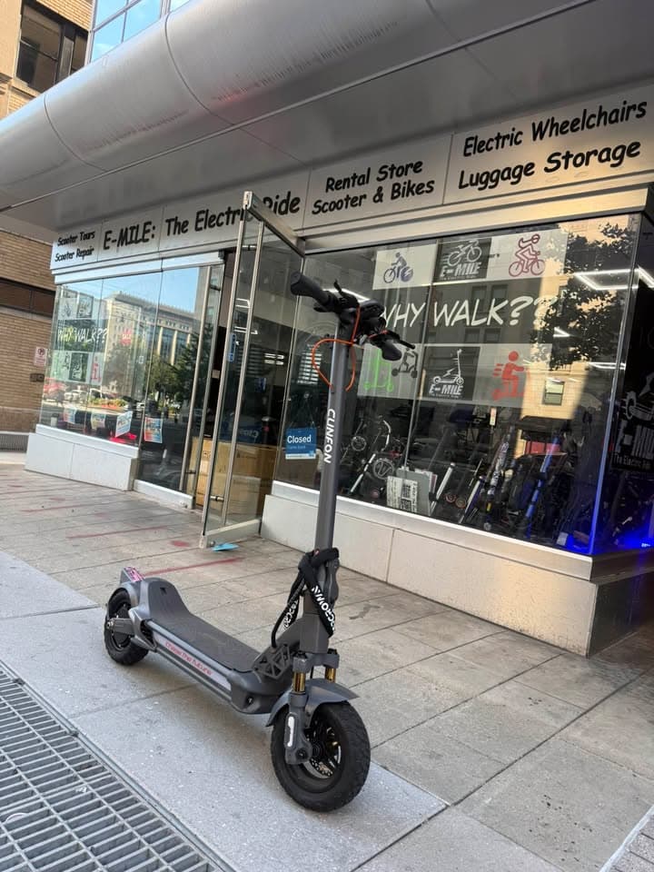 Grey electric scooter parked on a sidewalk in front of an electric vehicle rental store.