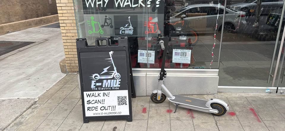 Silver electric scooter parked on a sidewalk next to a black E-MILE rental sign.