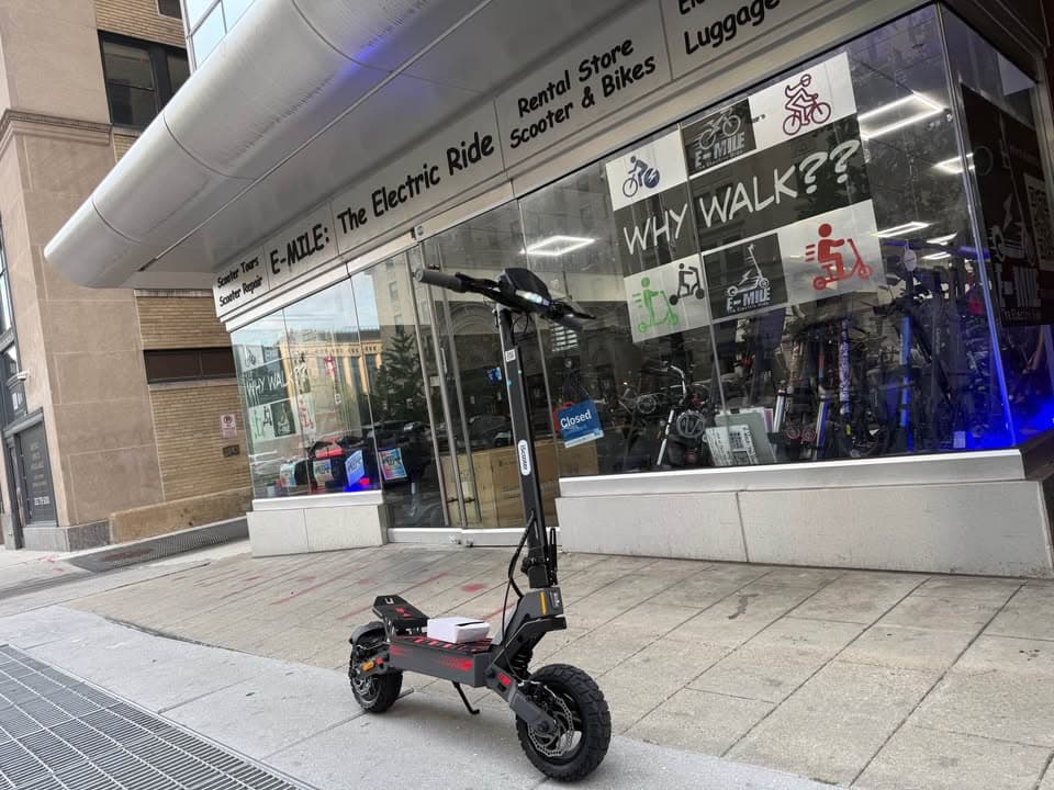 Black electric scooter with off-road tires parked on a sidewalk outside an E-MILE shop.