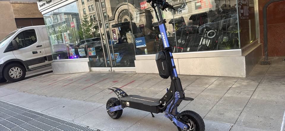 Blue electric scooter with a black storage bag parked on a sidewalk outside a storefront.