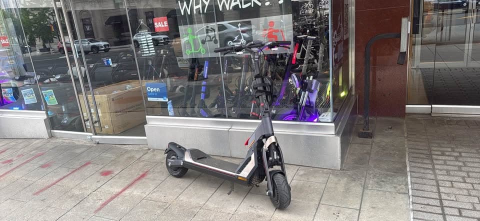 Black electric scooter parked on a sidewalk in front of a shop's glass window.